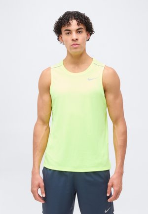 Hombre atlético con cabello rizado que lleva una camiseta deportiva sin mangas de color amarillo neón y pantalones cortos azul oscuro, de pie contra un fondo claro y liso.