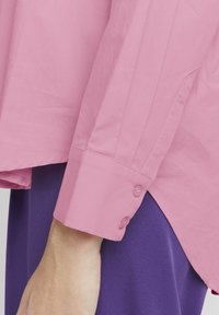 Rosa Hemd mit Knopfleiste, aus glattem Material, langen Ärmeln und geradem Schnitt. Verfügt über zwei Knöpfe an den Manschetten und einen Seitenschlitz.