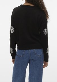 Svart stickad tröja med långa ärmar, prydd med vita snöflingemönster på armbågarna. Matchad med ljusblå vida jeans.