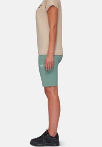Lys beige kortærmet t-shirt parret med teal shorts, med et logo, og sorte sportssko med textured detaljer.