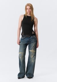 Haut noir sans manches avec laçage sur les côtés et fentes, associé à un jean denim large déchiré avec des éclaboussures de peinture et des ourlets effilochés.