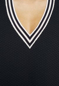 Navy texturovaný top s hlubokým V-výstřihem a pruhovaným lemem v bílé a námořnické barvě. Detailní pohled na výstřih a vzor látky.