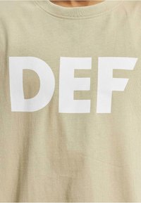 Lys beige T-skjorte med fete hvite bokstaver som skriver "DEF." Stoffet ser mykt ut med en glatt tekstur, og rundhalseringen er enkel.