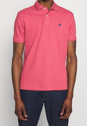 Mężczyzna ubrany w różową koszulkę polo z krótkim rękawem, z małym ciemnym logo na lewej piersi, w połączeniu z ciemnoniebieskimi dżinsami.