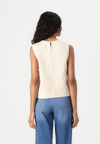 Top senza maniche di colore crema con una finitura testurizzata. Perle decorative adornano il retro, dotato di una zip centrale. Abbinato a jeans larghi blu.