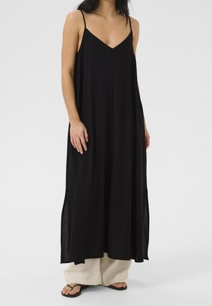 Kobieta w luźnej, czarnej, bezrękawnikowej, długiej sukience maxi z bocznymi rozcięciami, beżowe spodnie o szerokich nogawkach pod spodem oraz czarne sandały typu japonki.