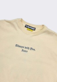 T-shirt beige a collo rotondo con testo gotico nero "Sempre con te. Iuter" centrato sotto il collo e etichetta nera del marchio IUTER all'interno del colletto.