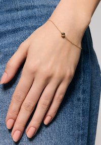 Złota bransoletka łańcuszkowa z okrągłą kulką, delikatny design, noszona na ręce na tle dżinsu. Gładka tekstura, minimalistyczny styl.