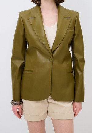 Femme portant un blazer en cuir vert olive sur un haut beige et un short beige retroussé, avec un bracelet à motif au poignet gauche.