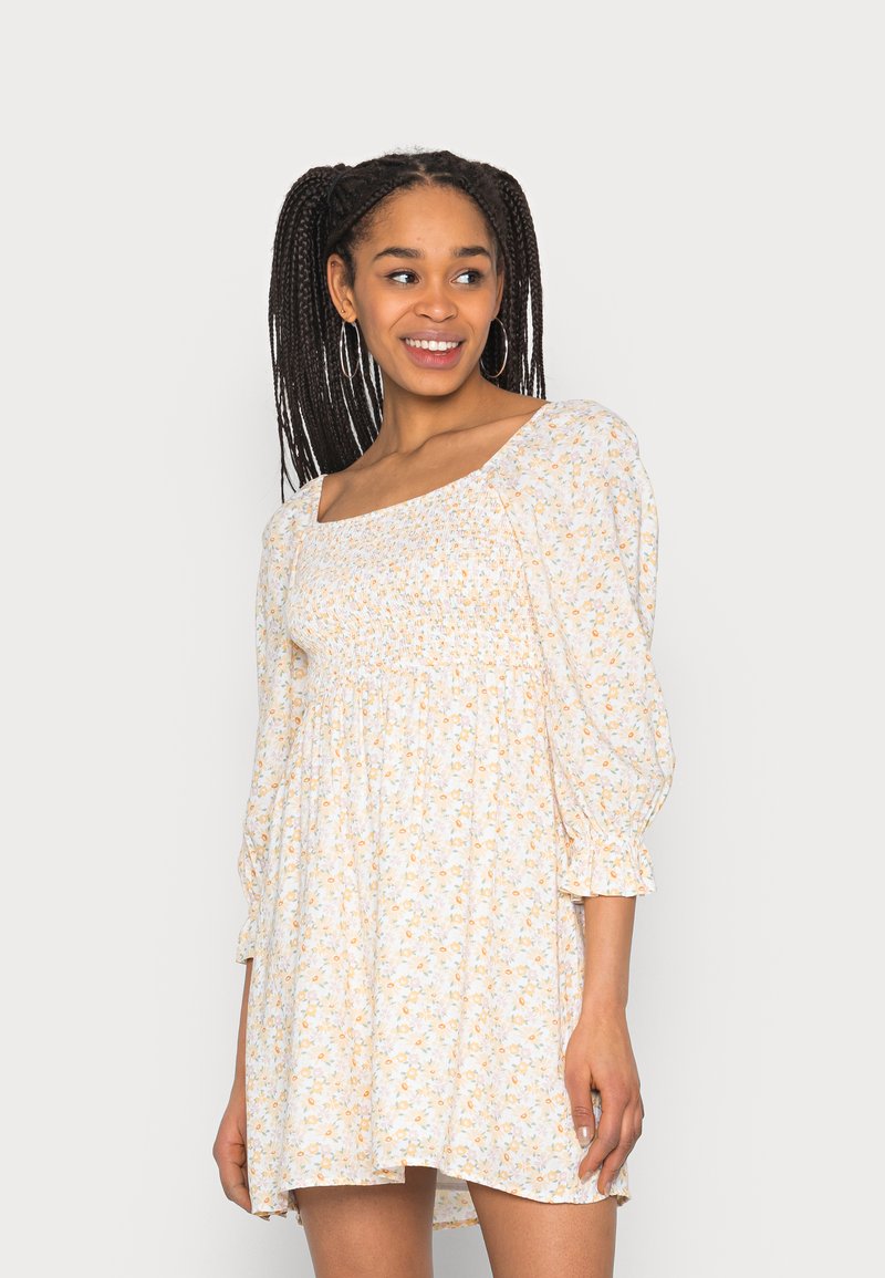 Vestido floral com decote quadrado, mangas bufantes e corpete franzido. O tecido leve apresenta pequenas flores laranja e azuis sobre um fundo branco.