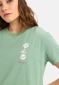 Lys grøn bomulds-t-shirt med et blomster- og bølgemotiv i hvid og pink, med ordet "Roxy" i kursiv skrift.