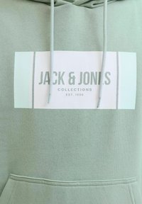 Lichtgroene hoodie met een voorzak en een wit rechthoekig logo met de tekst "JACK & JONES COLLECTIONS EST. 1990" en trekkoorden.