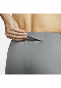 Mann hebt den Bund seiner grauen Sportshorts an, um eine kleine gefütterte Gesäßtasche mit Knopfverschluss zu zeigen.