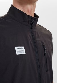 Schwarzige leichte Weste mit hohem Kragen, Frontreißverschluss und einer Brusttasche. Mit einem rechteckigen Markenlabel und glatter Textur.