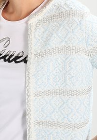 Ljusblå och vit stickad cardigan med ett geometriskt mönster i grå detaljer, öppen framsida och textilstruktur.