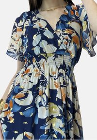 Marine sininen kukkamekko, jossa on V-mallinen kaula-aukko, lyhyet liehuvat hihat, kootut vyötärö ja kultaiset yksityiskohdat kevyessä kankaassa, jossa on eläväinen kuosi.