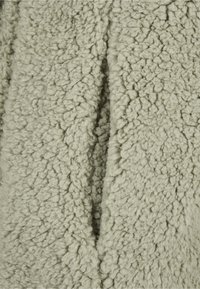 Nærbilde av lyst grått teksturert fleece-stoff med myke, tette og uregelmessige ull-lignende fibre som danner en myk overflate.