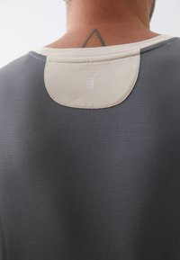 Grijs sportshirt met gestructureerde stof, contrasterende beige halslijn en een klein logo op de rug. Close-up van het kraaggebied.
