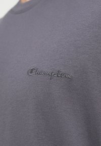 Hallhalli puuvillane t-särk, millel on rindkere peenelt tikitud "Champion" logo. Kangas on sileda tekstuuriga ja klassikalise ümmarguse kaelusega disain.