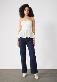 Béžový peplum top s tenkými ramínky, zkombinovaný s tmavomodrými širokými džínami a světle barevnými botami s špičatou špičkou. Hladká látka a vrstvený design.