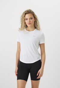 Camisa deportiva blanca de manga corta con cuello redondo y logo de Nike. Combinada con pantalones cortos negros ajustados que presentan detalles de costura lateral.