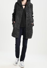 Grå, texturerad lång kappa med huva, med framknäppningar och sidofickor, tillsammans med svart topp och marinblå byxor, samt silverfärgade sneakers.