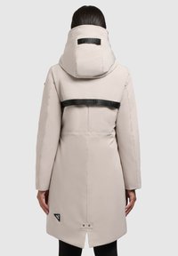 Vrouw in een lichtbeige lange winterjas met capuchon en een zwarte merkstrop over de bovenrug, gezien vanaf de achterkant.