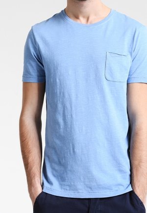 Mand iført en lyseblå t-shirt med korte ærmer og en brystlomme på venstre side, hænderne i lommerne på mørke bukser, uden synligt hoved eller baggrundsdetaljer.