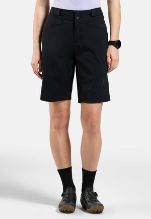 Schwarze Shorts mit glatter Textur, die zwei frontale Taschen und ein Markenlogo auf dem Oberschenkel aufweisen, kombiniert mit schwarzen Fahrradschoenen und Socken.