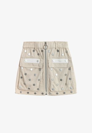 Minifalda beige con cremallera frontal, dos grandes bolsillos con solapa, cinturilla elástica y tachuelas circulares plateadas dispersas por la tela.