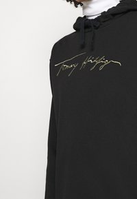 Černá bavlněná mikina s kapucí a měkkou texturou. Má kapuci se stahovací šňůrkou a zlatě vyšívané logo "Tommy Hilfiger" na hrudi.