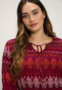 Rote Bluse mit Muster, rundem Ausschnitt und dekorativem Bindeband. Sie weist komplizierte geometrische Designs in Orange, Weiß und Schwarz auf.