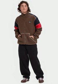 Mladic stoji s rukama u džepovima, odjeven u smeđu flis jaknu s crvenim i mornarsko plavim rukavima, crne hlače i smeđe tenisice.