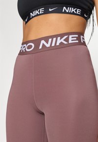 Nike Pro retuus pehmest lilakalt värvil, millel on lai elastne vöökoht, millel on valged "NIKE PRO" kirjad. Sile ja veniv kangas.