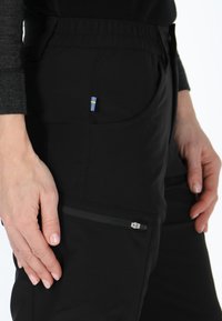 Svarta byxor i stretchmaterial med en sidoficka med dragkedja och en liten svensk flagga som accent på framfickan.