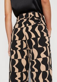 Vida byxorna med ett abstrakt svart och beige vågmönster. De har hög midja, subtila fickor och en mjuk textilfinish.