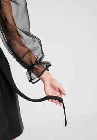 Svart klänning med genomskinliga puffärmar. Ärmarna har elastiska cuffs och en mjuk, flödande textur. Ett långt svart band hänger från midjan.