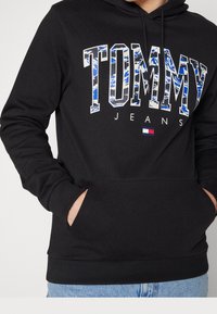 Sudadera negra con bolsillo canguro en la parte delantera, con el gráfico "TOMMY JEANS" en azul y blanco; puños y dobladillo acanalados para un ajuste ceñido.