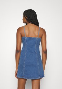 Denim dress in blue, fitted with thin straps. Features a straight cut, back button detail, and a curved hem. Smooth texture.