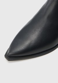 Μαύρο δερμάτινο μποτάκι αστραγάλου με μυτερή μύτη, επίπεδη σόλα και λεία υφή. Ο σχεδια�σμός διαθέτει κομψό προφίλ με γωνιώδεις λεπτομέρειες.