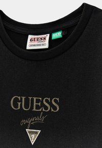 Czarna bawełniana koszulka z okrągłym dekoltem. Posiada złote logo "GUESS" oraz napis "originals", a także srebrny trójkątny akcent z czarnym logo.