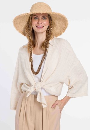 Linen wrap top in beige, tied at the waist. Paired with a white shirt and wide beige trousers. Features a wide-brim straw hat and chunky necklace.