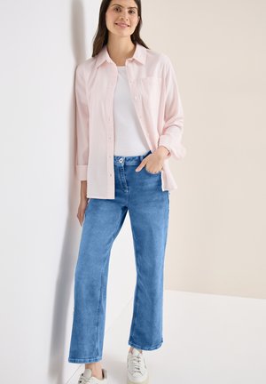 Jeune femme souriante, portant une chemise rose clair boutonnée sur un haut blanc, un jean bleu et des baskets blanches, debout contre un mur d'angle.