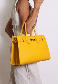 Donna che indossa un vestito senza maniche bianco tiene una grande borsa in pelle gialla con doppio manico e chiusura dorata all'altezza del gomito.
