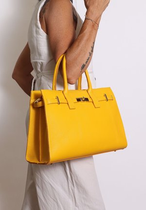 Femme portant une robe sans manches blanche tient un grand sac à main en cuir jaune avec des doubles poignées et un fermoir doré au niveau du coude.