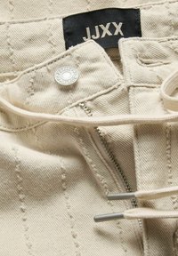 Gros plan sur un pantalon en jean beige avec fermeture éclair, bouton métallique gravé "JJXX", cordon de serrage et étiquette en tissu noire avec l'inscription "JJXX".