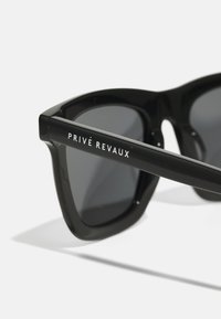 Black rectangular sunglasses with dark lenses. Features a thick frame and temples. “PRIVÉ REVAUX” logo in white on the side. Glossy finish.