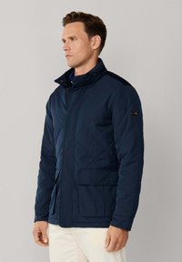 Marineblauwe doorgestikte jas met een hoge kraag, voorzien van een ritssluiting en zijzakken. Gemaakt van een glad, lichtgewicht materiaal.