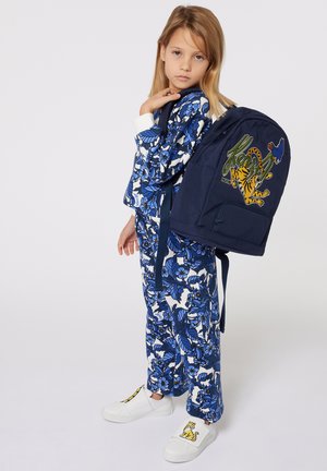 Marineblauw stoffen backpack met een ontwerp van een tijger en een vogel; bloemenpatroon outfit in blauw en wit; witte schoenen met een gele accent.