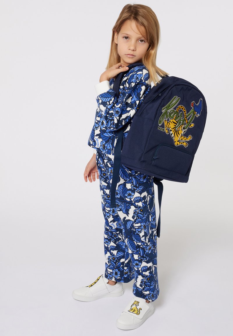 Sac à dos en tissu bleu marine avec un design de tigre et d'oiseau ; tenue à motifs floraux en bleu et blanc ; chaussures blanches avec un accent jaune.